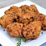 A white square plate filled with pieces of golden-brown fried chicken, garnished with small sprigs of fresh herbs.