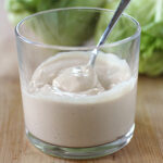A clear glass filled with creamy salad dressing, with a spoon inside. The glass is on a wooden surface, and leafy green lettuce is blurred in the background.