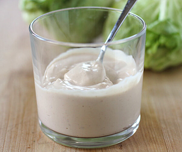 A clear glass filled with creamy salad dressing, with a spoon inside. The glass is on a wooden surface, and leafy green lettuce is blurred in the background.