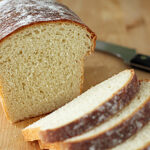 A loaf of homemade bread on a wooden surface with three slices cut, next to a bread knife with a black handle. The bread has a golden-brown crust and a soft, airy interior.