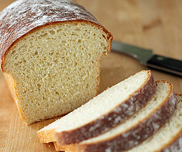 A loaf of homemade bread on a wooden surface with three slices cut, next to a bread knife with a black handle. The bread has a golden-brown crust and a soft, airy interior.