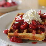 Two golden waffles topped with sliced strawberries, strawberry syrup, and a dollop of whipped cream are served on a white plate. A glass of orange juice and a bowl are blurred in the background.