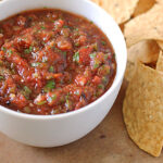 A white bowl filled with chunky red salsa garnished with herbs sits on a wooden surface next to a pile of crispy tortilla chips.