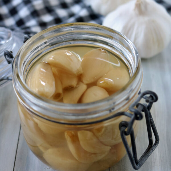 An open glass jar filled with peeled garlic cloves in liquid sits on a light wooden surface, with whole garlic bulbs and a black-and-white checkered cloth in the background.
