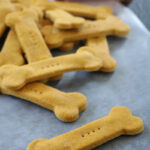 Bone-shaped dog biscuits are arranged on a sheet of parchment paper, with a bone-shaped cookie cutter in the background. The biscuits are golden brown and have small holes pricked in the center.