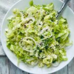 A white bowl filled with Crunchy Romaine Salad—chopped romaine lettuce, thinly sliced onions, grated parmesan cheese, and seasoning—placed on a rustic wooden table with a white napkin and a fork.