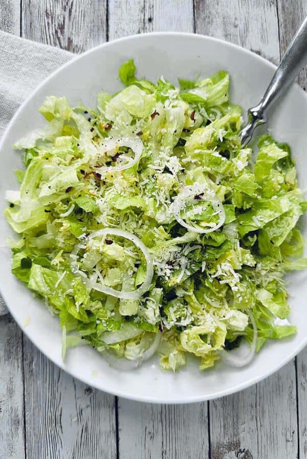 A white plate with a fresh Crunchy Romaine Salad, featuring chopped lettuce, thinly sliced onions, grated Parmesan cheese, and a sprinkle of seasoning, on a rustic wooden table with a silver fork.