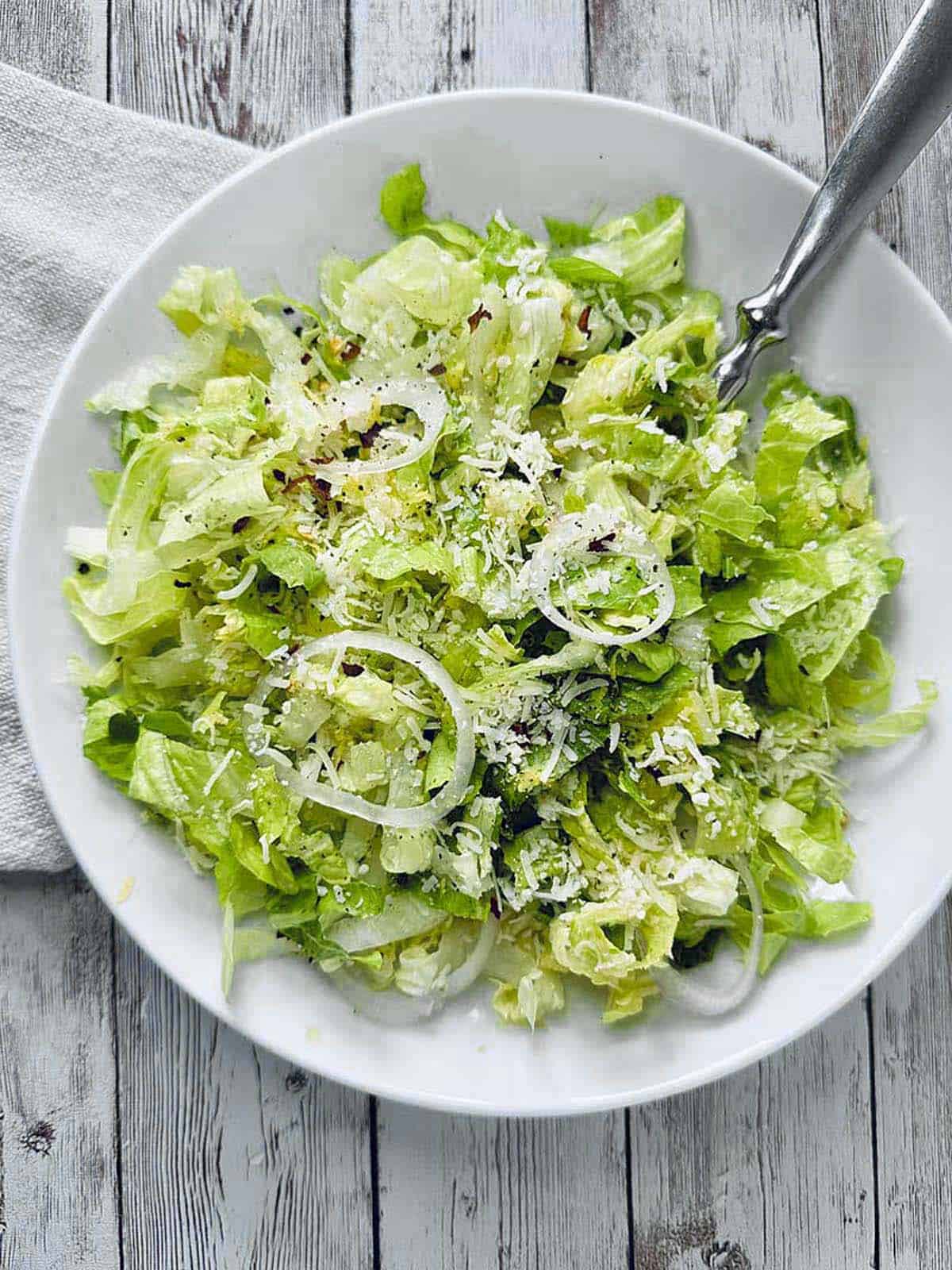 A white plate with a fresh Crunchy Romaine Salad, featuring chopped lettuce, thinly sliced onions, grated Parmesan cheese, and a sprinkle of seasoning, on a rustic wooden table with a silver fork.