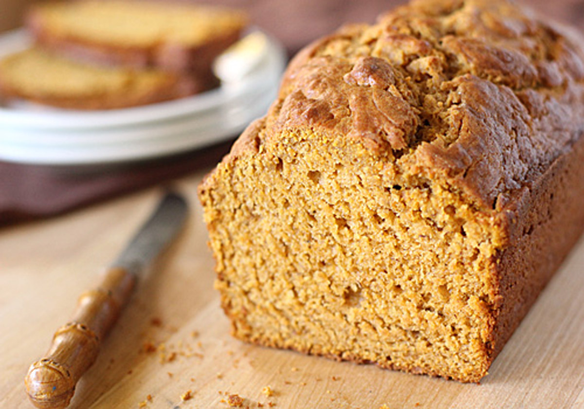 A close-up of a loaf of pumpkin bread on a wooden surface, with a knife beside it and a plate of sliced bread in the background. The bread has a golden-brown crust and moist, textured interior.