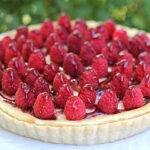 A tart topped with fresh raspberries is arranged on a white plate. The tart has a golden crust, creamy filling, and is glazed for a glossy finish. Green blurred background suggests the image was taken outdoors.