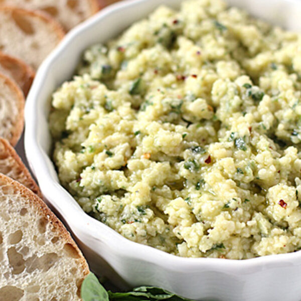 A white bowl filled with a creamy, herbed ricotta cheese spread, surrounded by several slices of crusty baguette bread. Fresh basil leaves are placed beside the bowl.