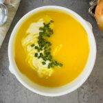A bowl of creamy yellow soup topped with a swirl of cream and chopped chives, placed on a gray surface next to a piece of bread.