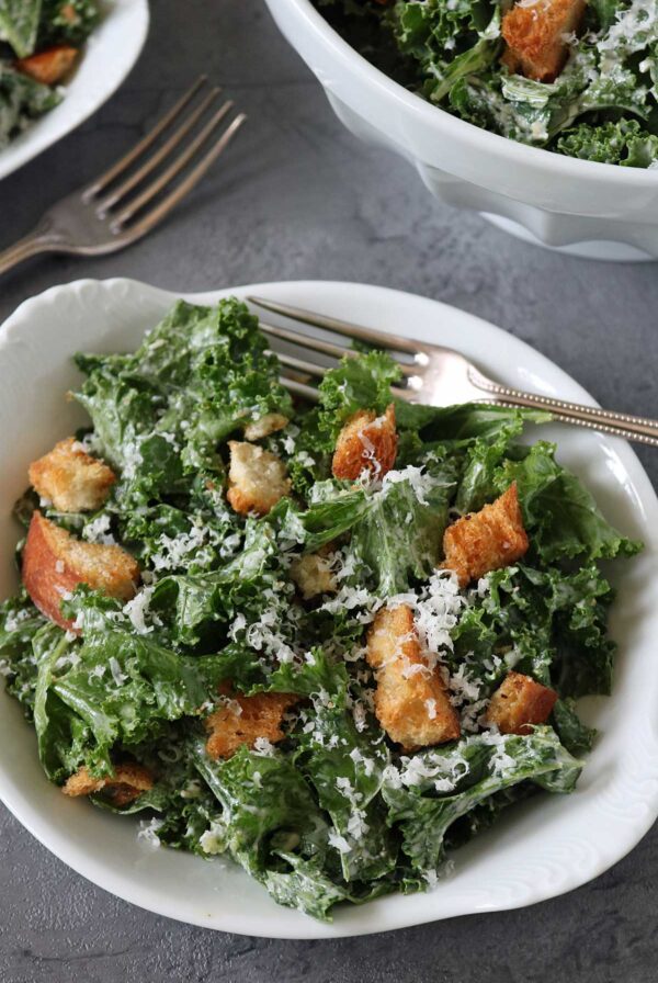 A white bowl filled with kale Caesar salad topped with croutons and grated cheese, with a fork resting inside. Another bowl and a fork are visible in the background on a gray surface.