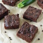 Squares of chocolate zucchini brownies are arranged on parchment paper, with a fresh zucchini and scattered chocolate chips nearby. A knife rests at the bottom of the image.