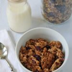 A white bowl of granola sits next to a glass jar of milk, a metal spoon, and a large jar of granola, all placed on a white surface with a folded white napkin.