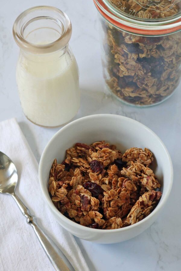 A white bowl of granola sits next to a glass jar of milk, a metal spoon, and a large jar of granola, all placed on a white surface with a folded white napkin.