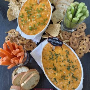 Two oval dishes of buffalo chicken dip, one with ranch and one with blue cheese, are surrounded by chips, pretzel crisps, sliced baguette, celery, and carrot sticks on a dark table. Small chalk signs label each dip.