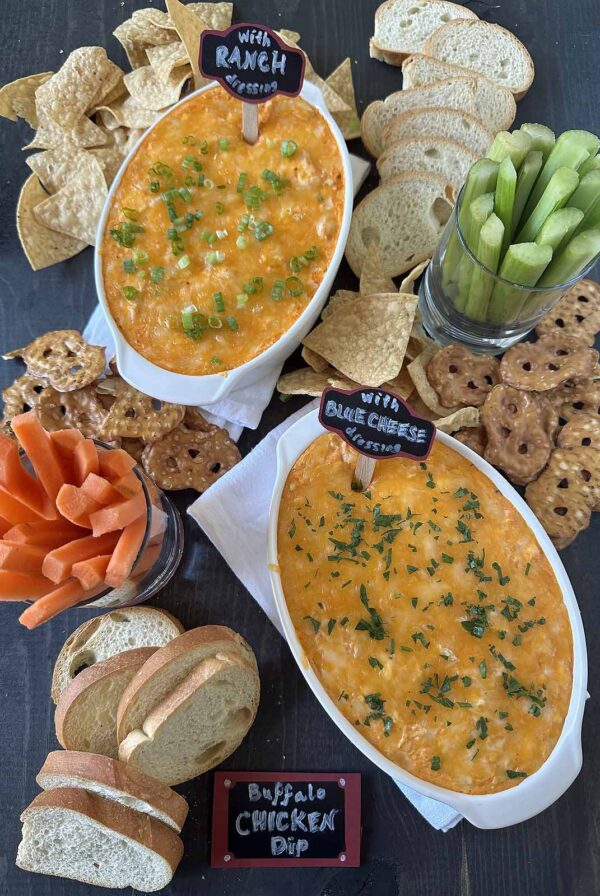 Two oval dishes of buffalo chicken dip, one with ranch and one with blue cheese, are surrounded by chips, pretzel crisps, sliced baguette, celery, and carrot sticks on a dark table. Small chalk signs label each dip.