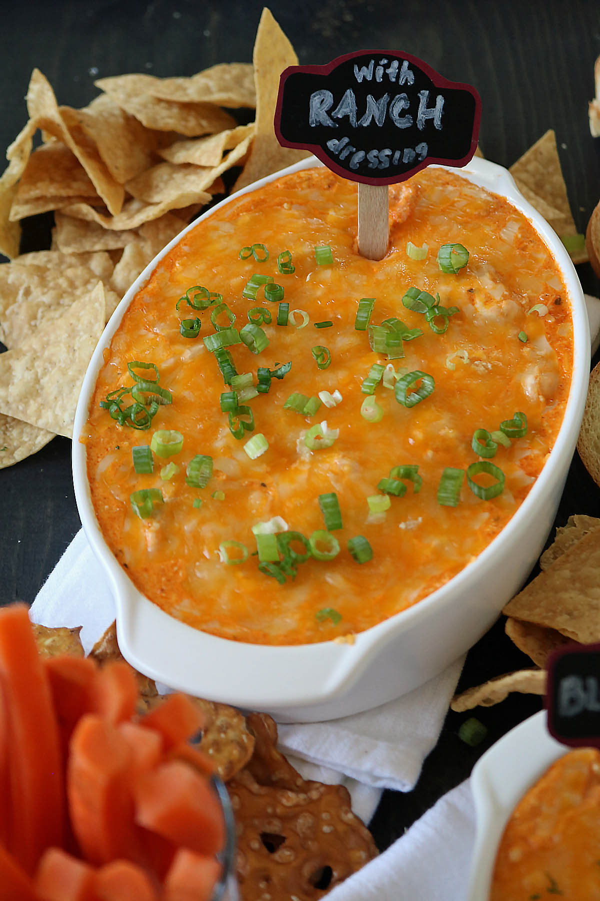 A white dish filled with cheesy, baked dip topped with chopped green onions. A small sign says With Ranch Dressing. Surrounding the dish are tortilla chips, carrot sticks, and pretzels.