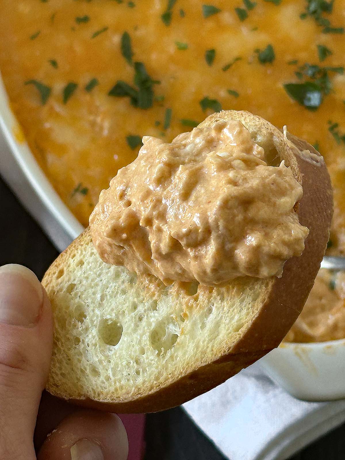 A hand holds a slice of bread topped with a creamy, orange dip. In the background, a dish filled with the same dip is garnished with chopped herbs.