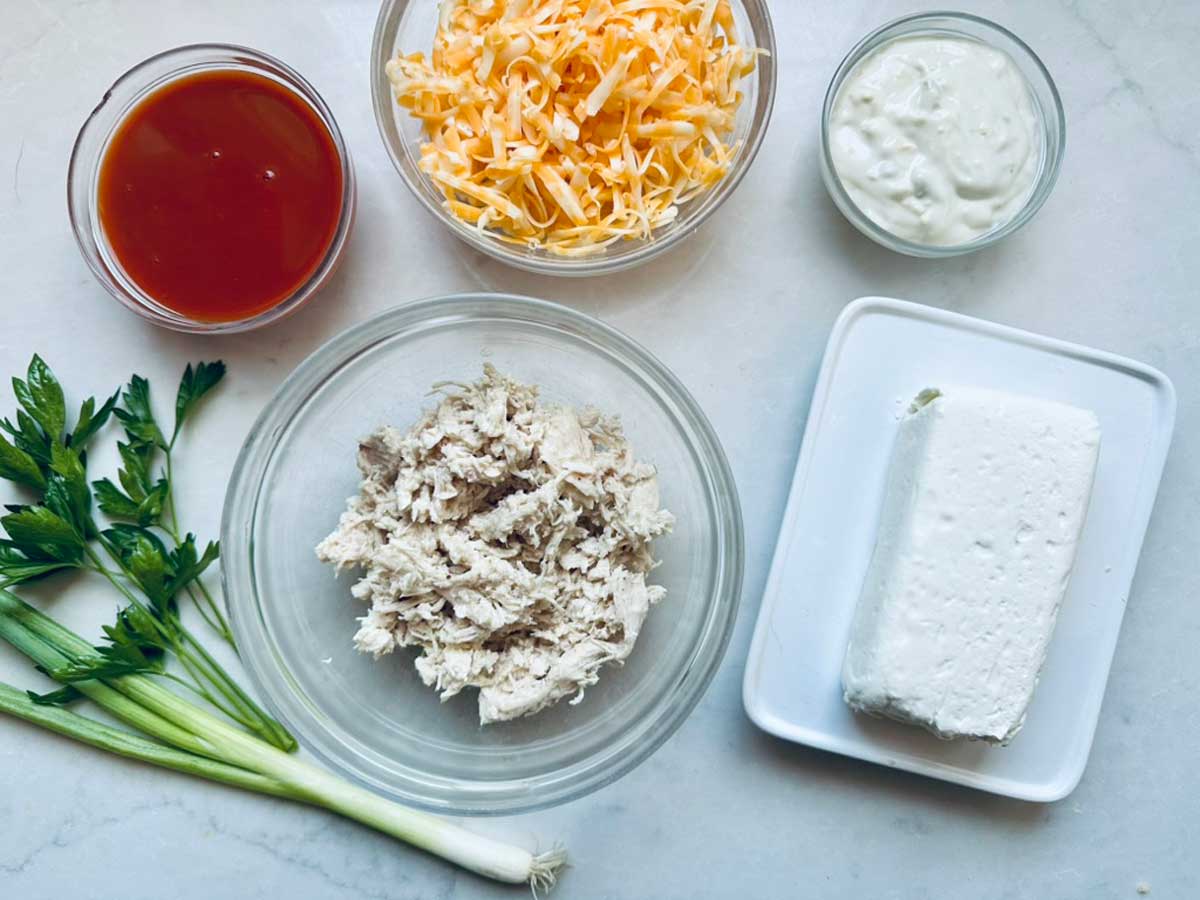A top-down view of ingredients on a white surface: a bowl of shredded chicken, grated cheese, buffalo sauce, ranch dressing, a block of cream cheese, green onions, and fresh parsley.