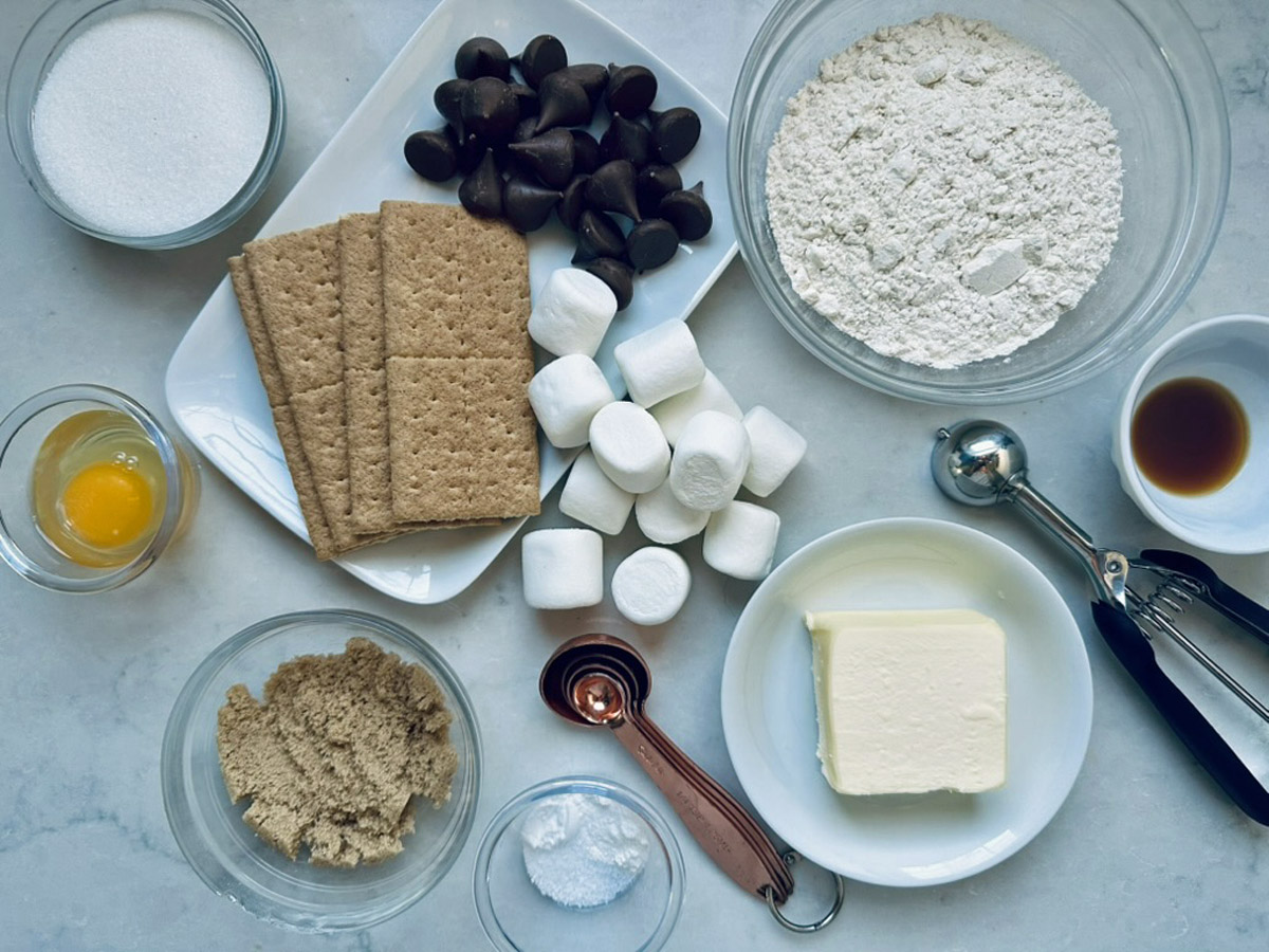 Baking ingredients on a white surface, including flour, sugar, brown sugar, butter, marshmallows, chocolate chips, graham crackers, an egg yolk, vanilla extract, baking soda, and measuring spoons.