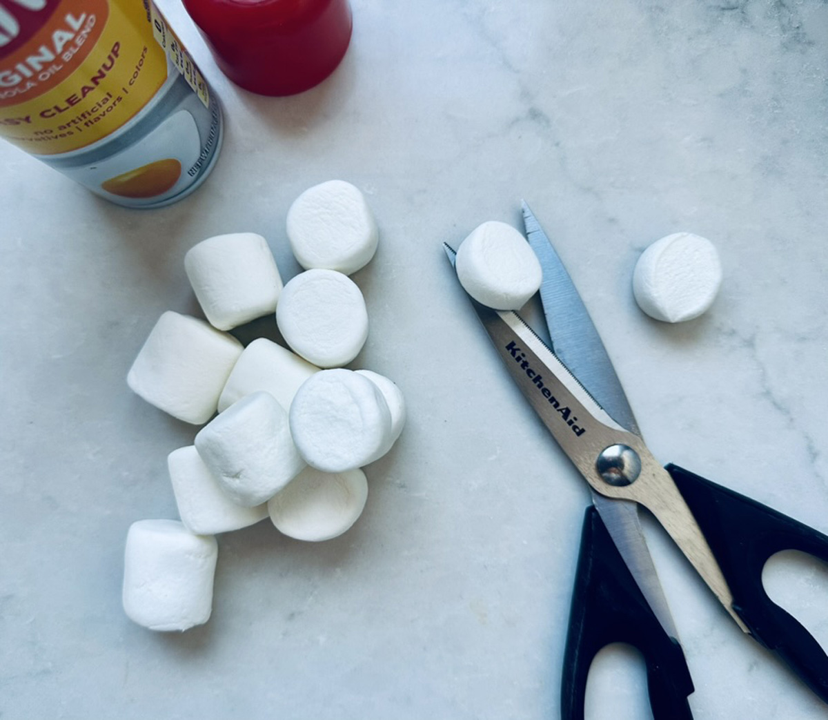 Large marshmallows on a marble surface, some whole and some sliced, with scissors labeled KitchenAid, a can of non-stick spray, and part of a red item visible at the top.