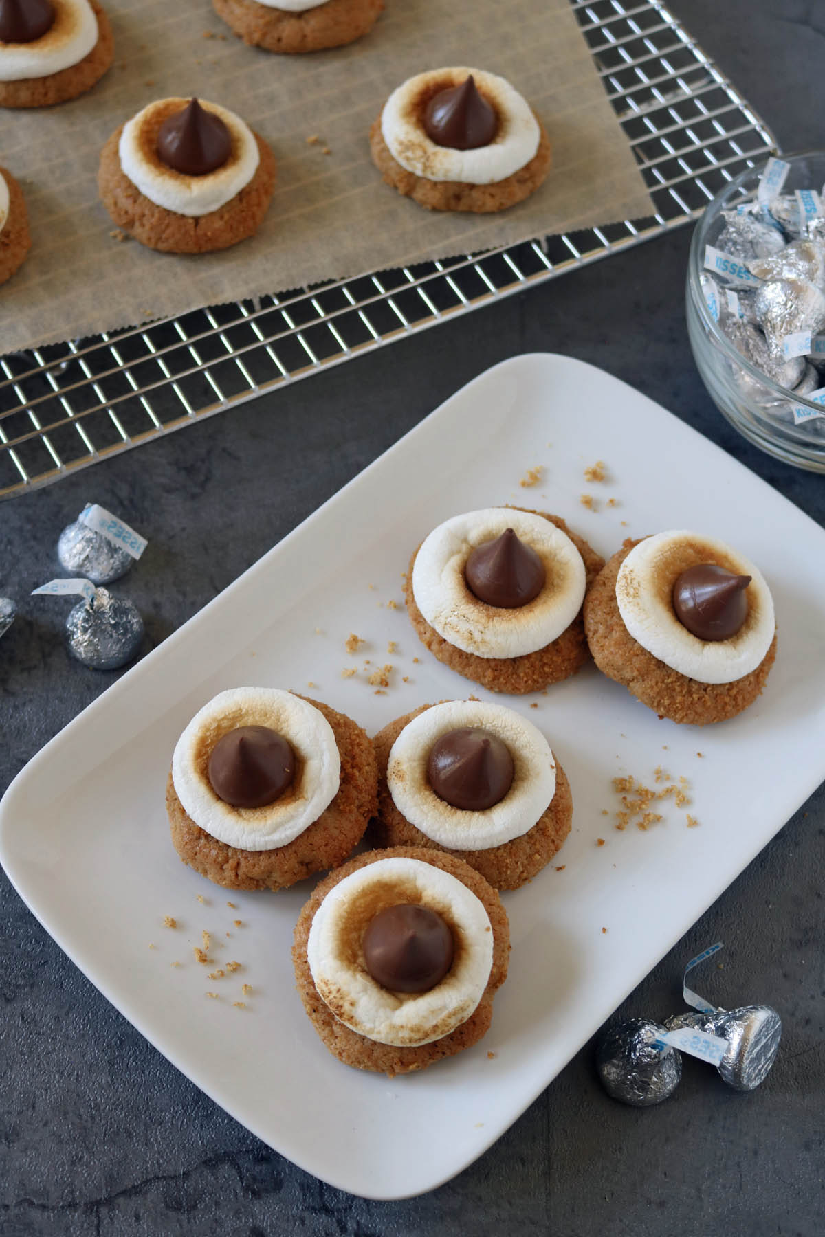Five cookies topped with marshmallow rings and chocolate kisses are arranged on a white plate, with more cookies on a cooling rack and wrapped chocolate kisses nearby on a gray surface.