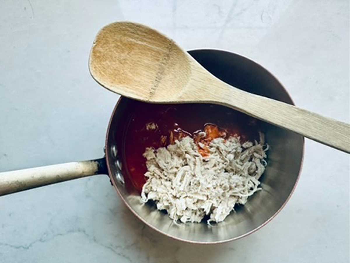 A saucepan with shredded chicken and red sauce inside, placed on a light countertop. A wooden spoon rests across the top of the saucepan.