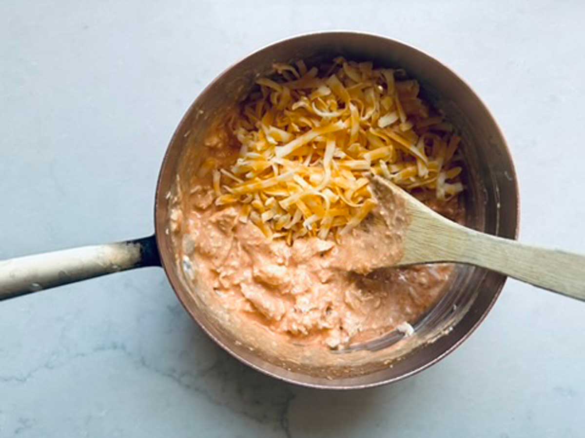 A pot filled with a creamy orange mixture topped with shredded cheese, being stirred with a wooden spoon, sits on a light-colored countertop.