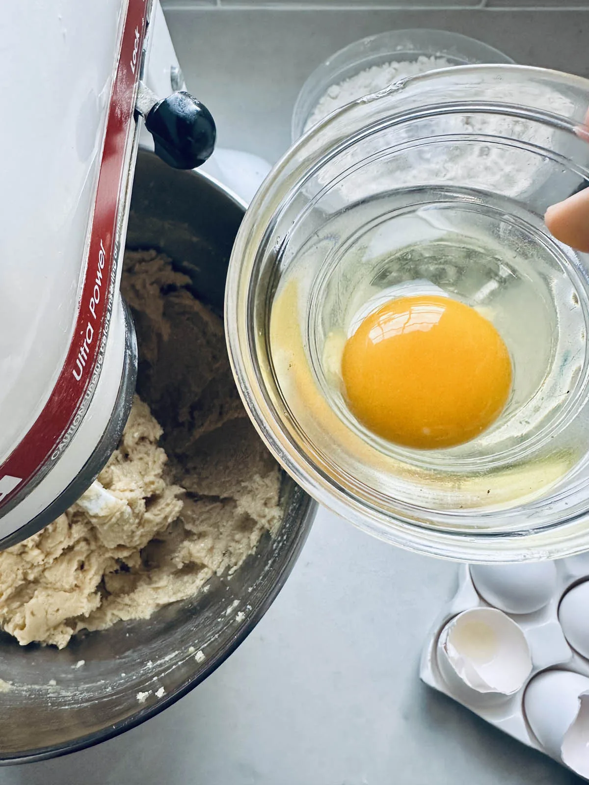 A hand holds a glass bowl with a cracked raw egg above a mixing bowl with dough in a stand mixer; an open carton of eggs is nearby on a light countertop.
