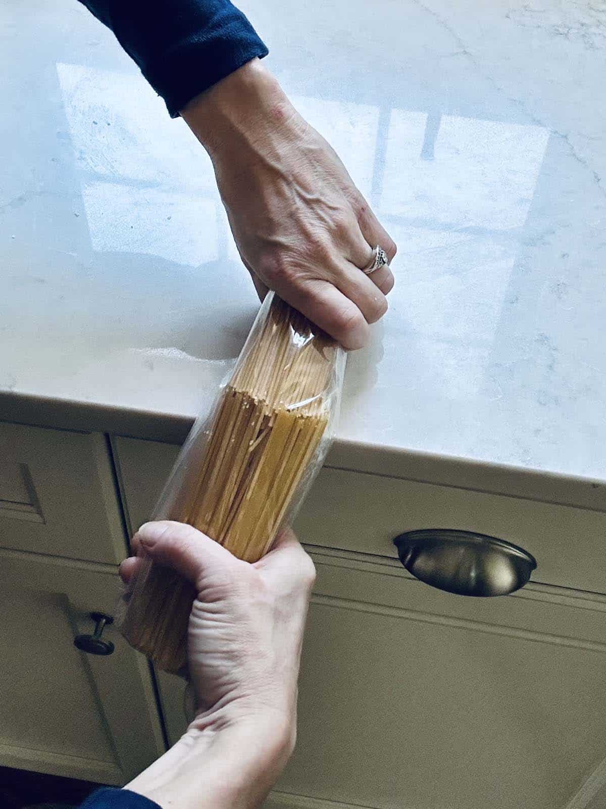 A person breaks a bundle of uncooked spaghetti in half over a kitchen counter, holding the pasta in both hands.
