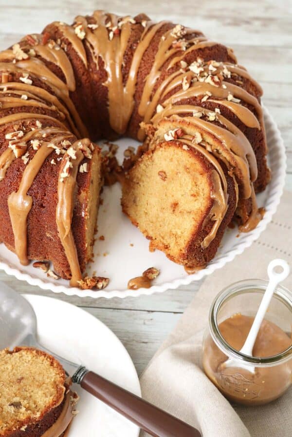 A bundt cake with caramel glaze and chopped pecans on top sits on a white plate. A slice has been cut and placed nearby, with a jar of caramel sauce and a spoon next to the cake.