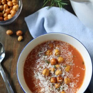 A bowl of tomato soup topped with crispy chickpeas and grated cheese, set on a wooden table beside a spoon, a blue napkin, a bowl of chickpeas, fresh rosemary, and a white pot.