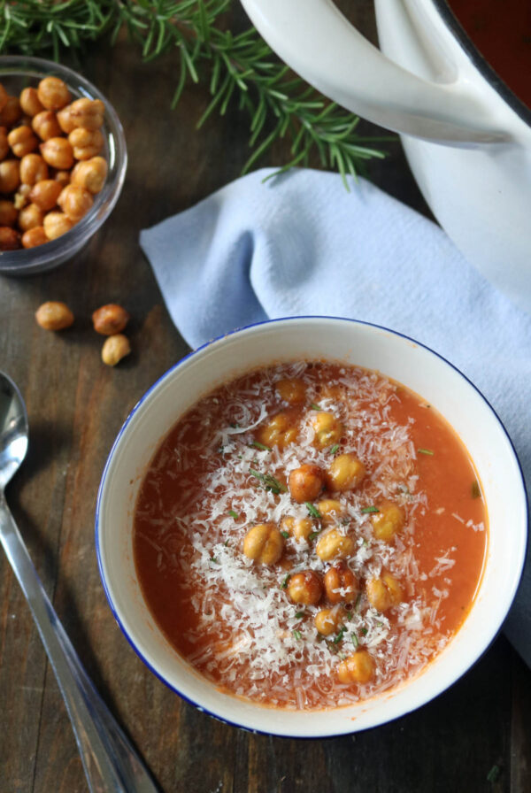 A bowl of tomato soup topped with crispy chickpeas and grated cheese, set on a wooden table beside a spoon, a blue napkin, a bowl of chickpeas, fresh rosemary, and a white pot.