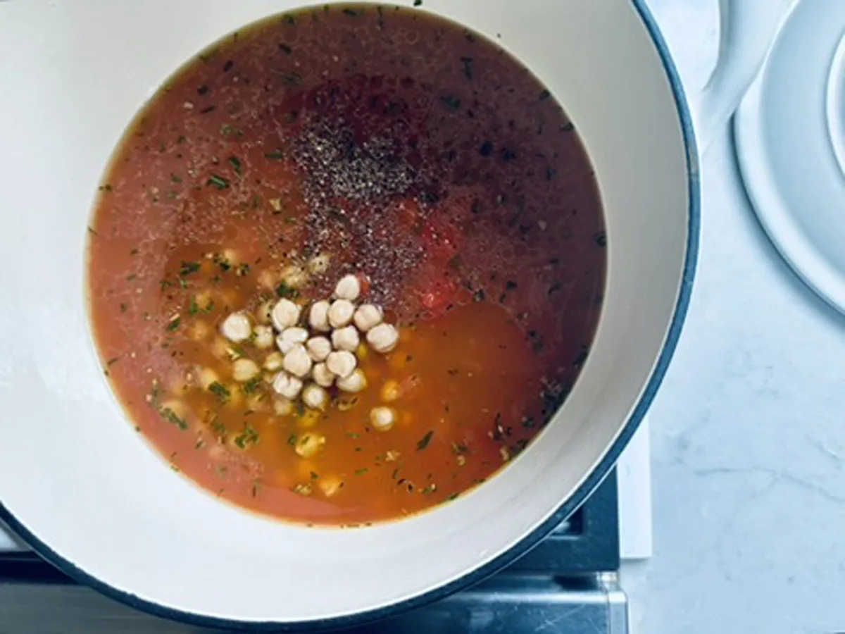 A white pot on a stove contains tomato-based soup with chickpeas, herbs, and ground pepper visible on the surface.
