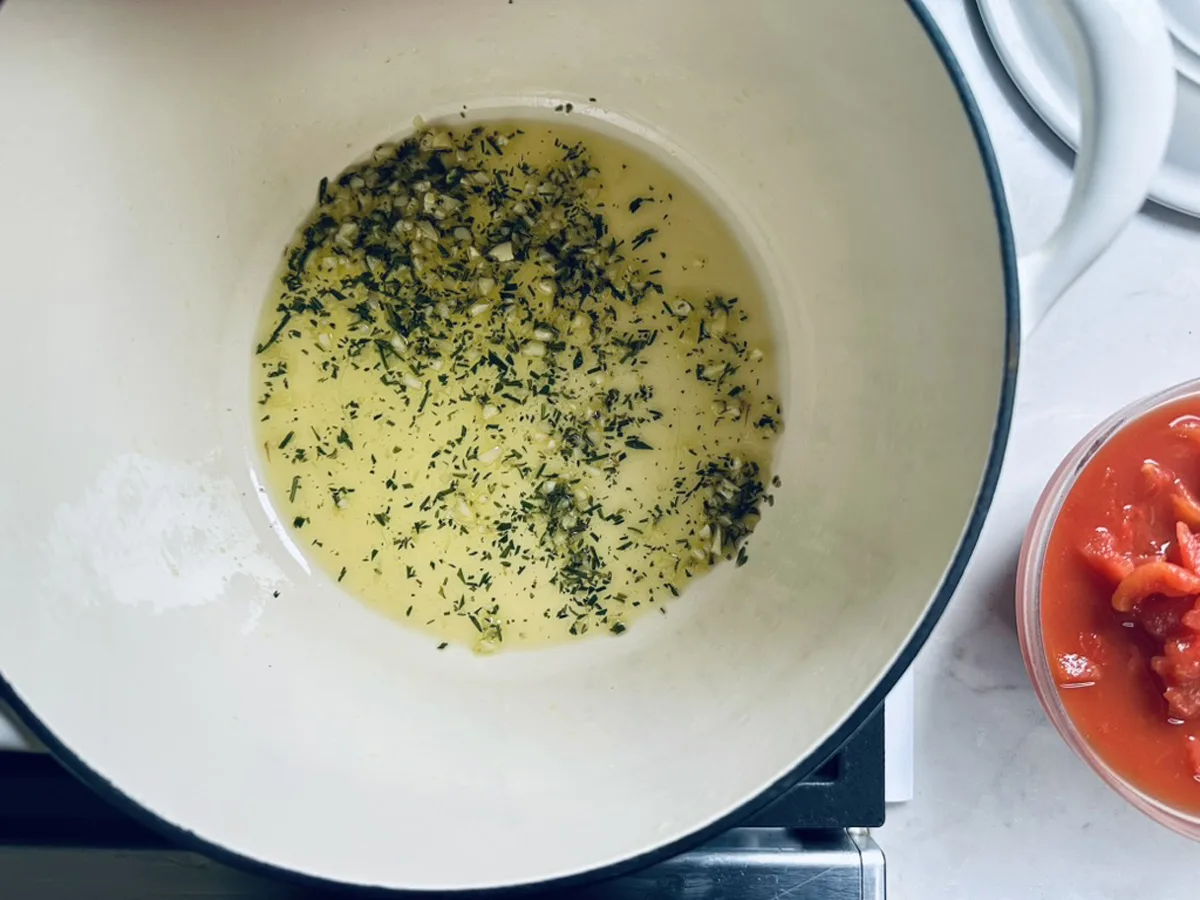 Chopped garlic and herbs saut&eacute;ing in oil in a white pot on a stovetop, with a bowl of diced tomatoes next to the pot.