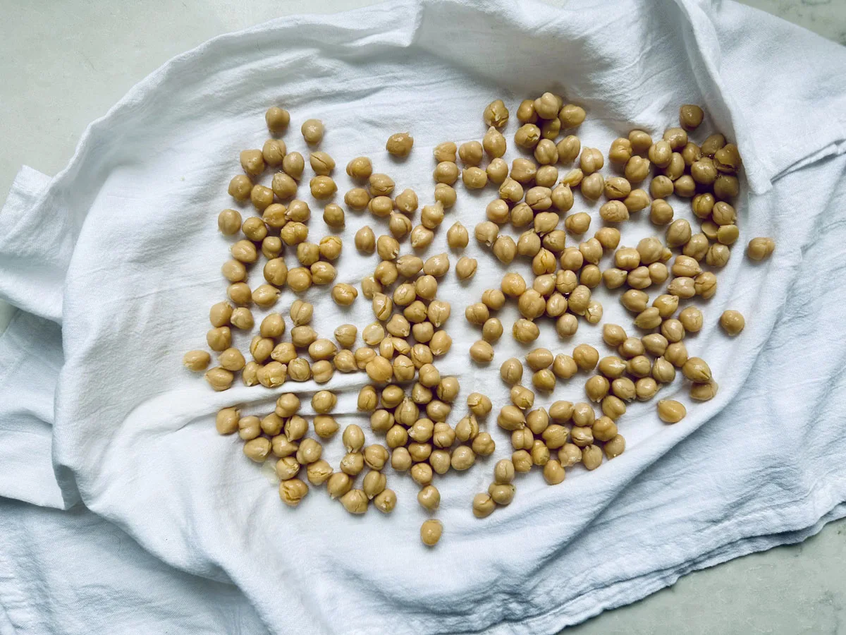 Cooked chickpeas spread out on a white cloth, drying in preparation for a roasted chickpeas recipe. The chickpeas are scattered in uneven clusters, with the slightly wrinkled cloth resting on a light-colored surface.