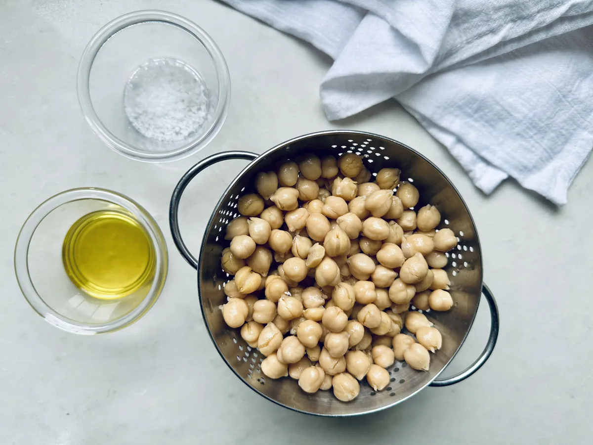 A metal colander filled with chickpeas sits on a light surface beside a small glass bowl of olive oil and a bowl of salt, ready for your roasted chickpeas recipe, with a white cloth in the corner.