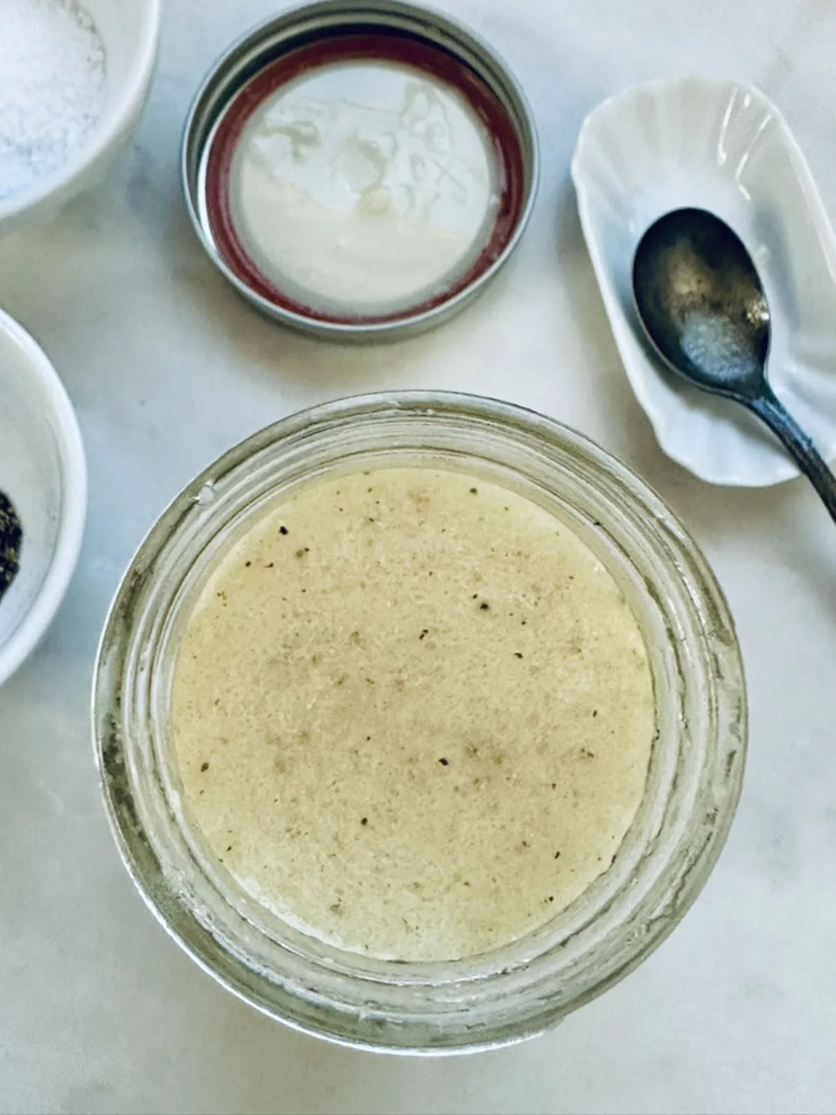 A jar filled with a creamy, speckled dressing is shown from above, next to an open lid, a spoon on a small dish, and bowls containing salt and pepper on a white surface.