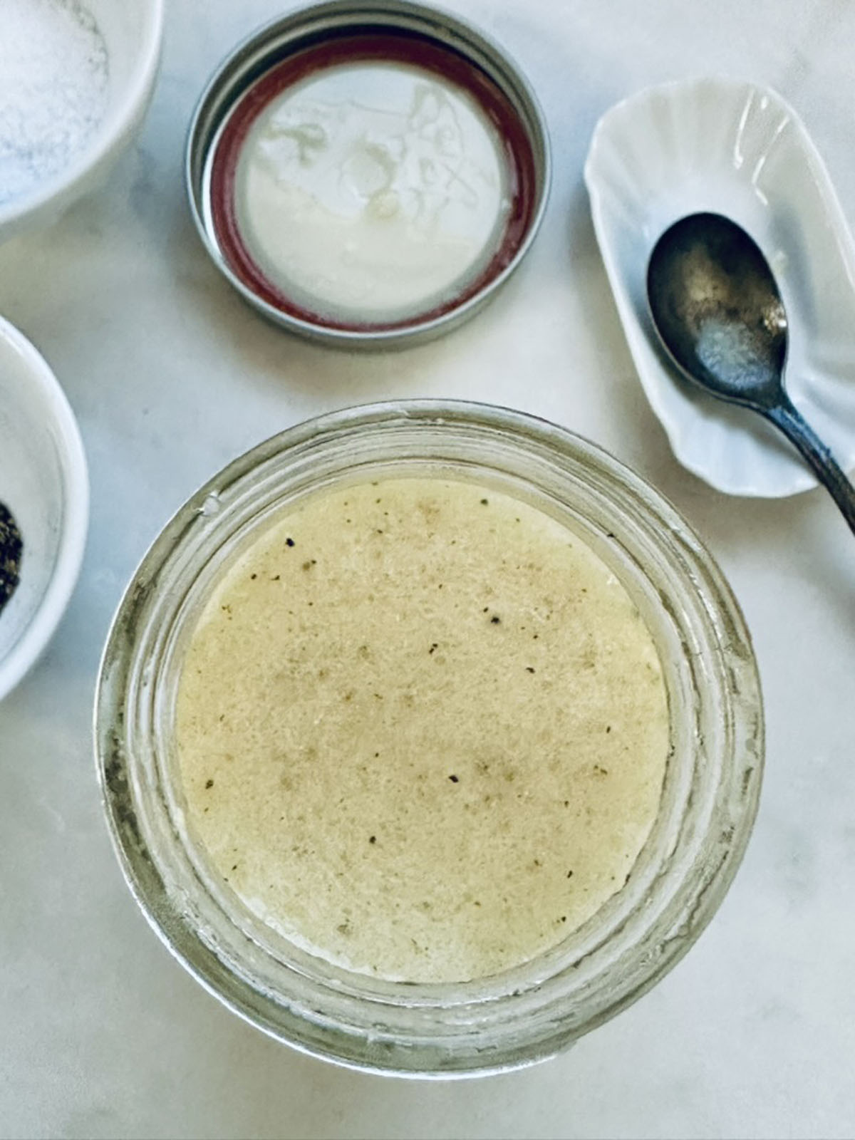 A jar filled with a creamy, speckled dressing is shown from above, next to an open lid, a spoon on a small dish, and bowls containing salt and pepper on a white surface.
