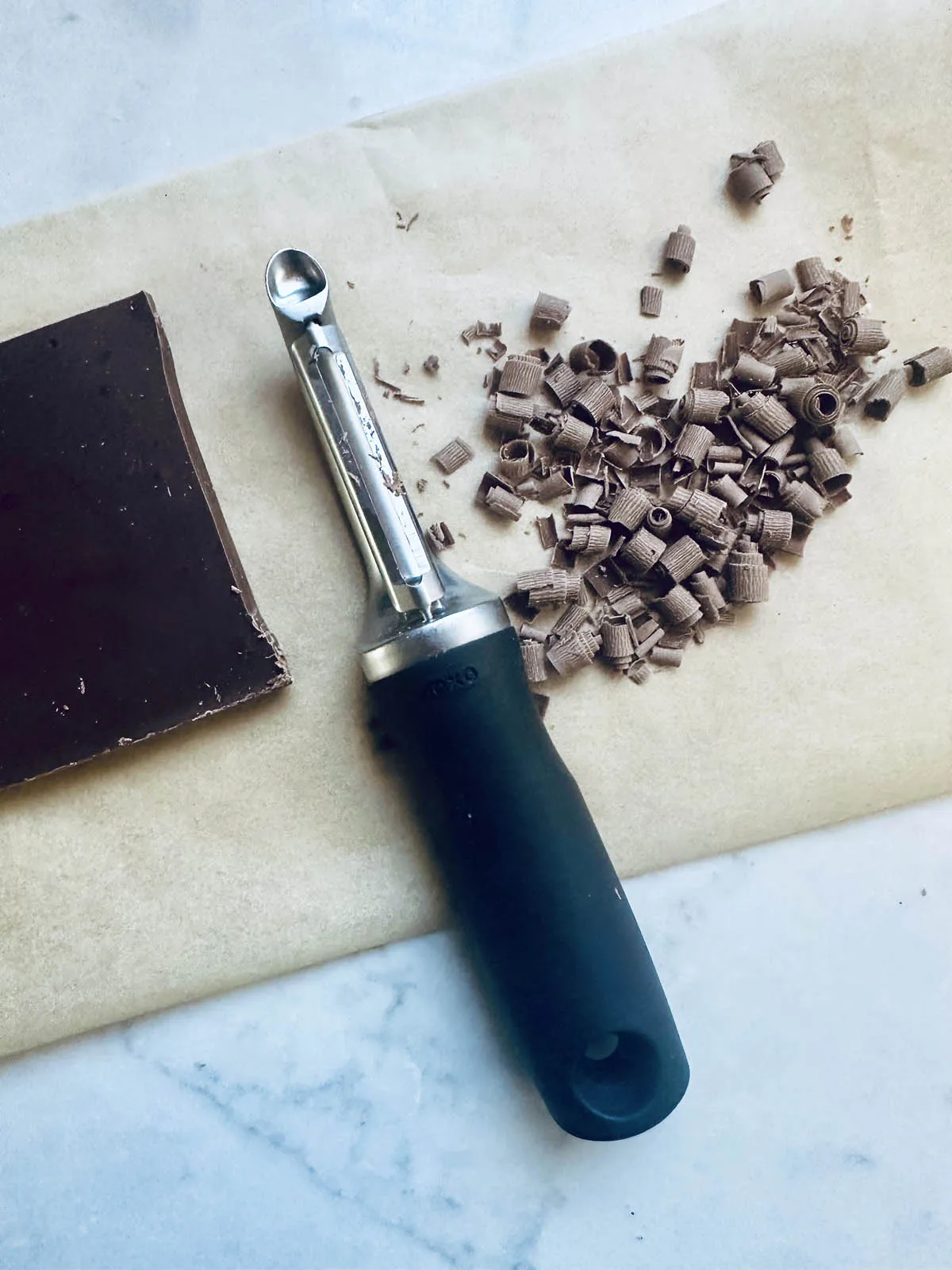 A vegetable peeler rests on parchment paper next to a block of chocolate and a small pile of chocolate curls—perfect for garnishing a decadent peanut butter chocolate tart.