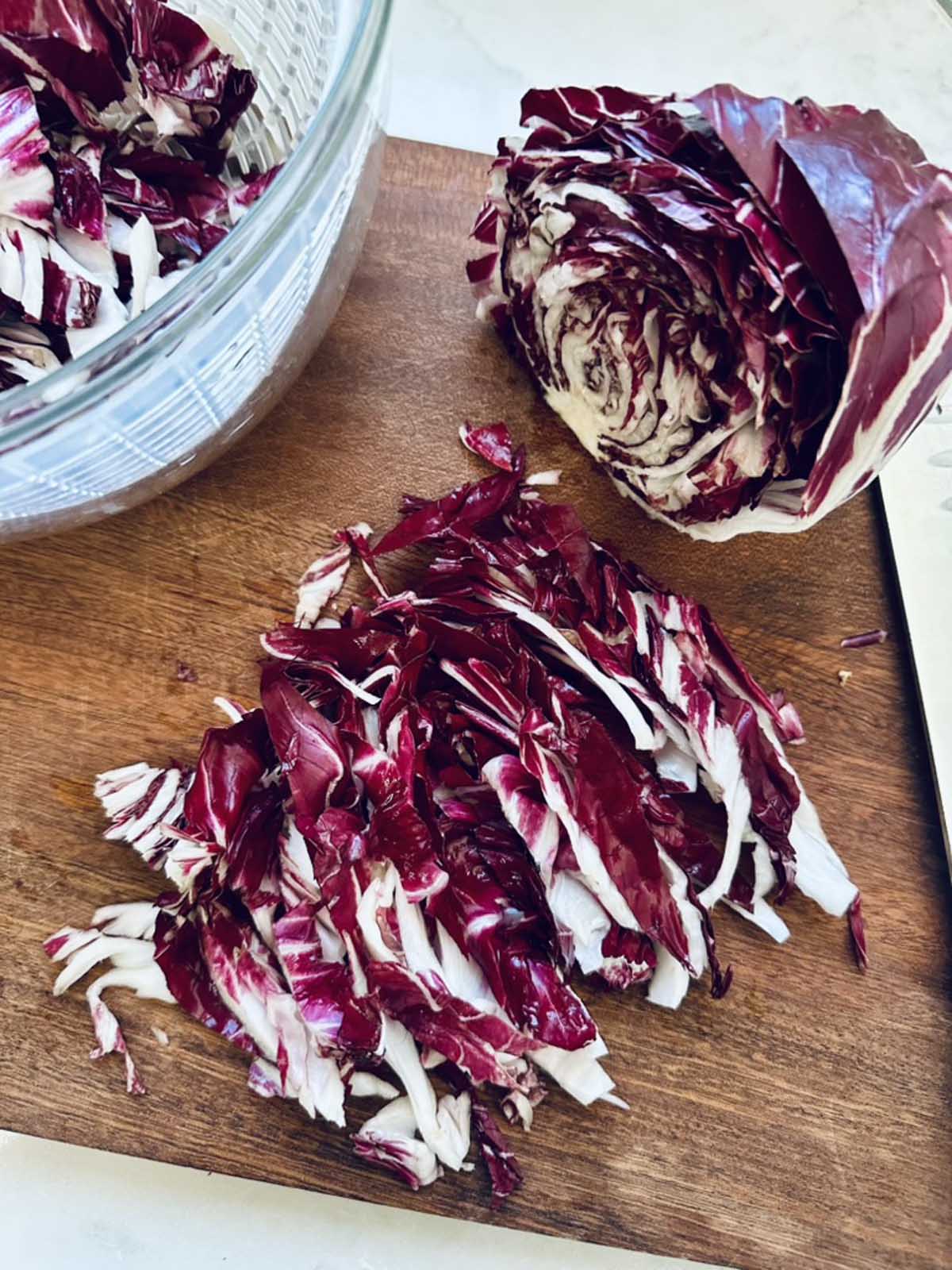 A halved head of radicchio and thinly sliced radicchio pieces are on a wooden cutting board, with a glass bowl nearby.