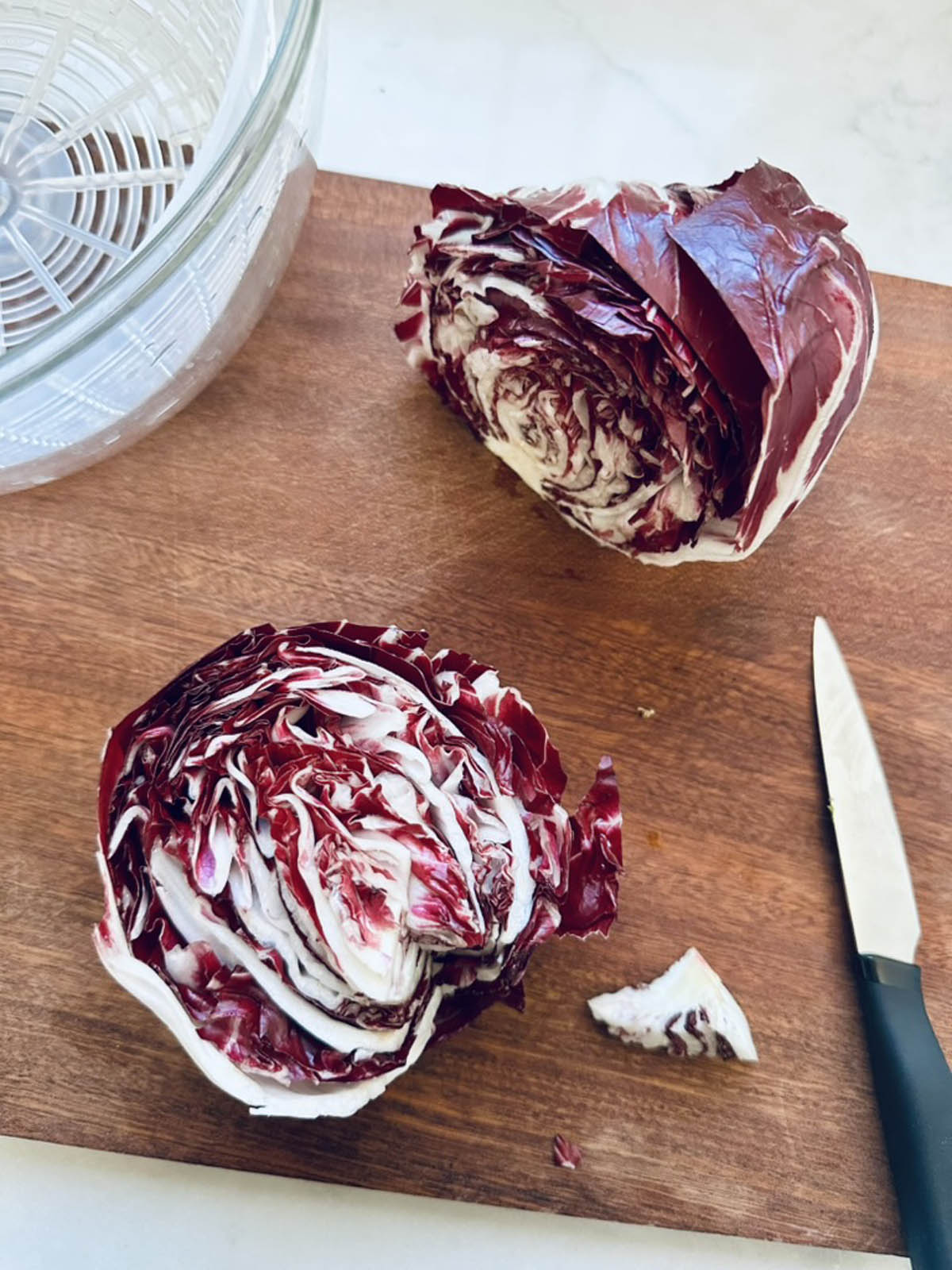 A head of radicchio cut in half sits on a wooden cutting board next to a small knife and a glass salad spinner.