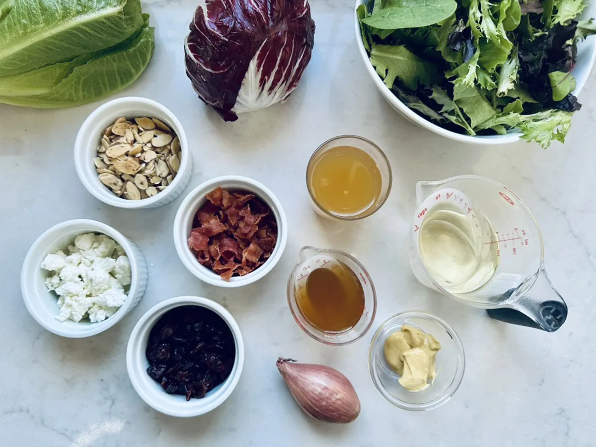 A variety of salad ingredients on a white surface, including leafy greens, radicchio, romaine, nuts, crumbled cheese, dried fruit, crispy bacon, a shallot, and small containers of vinaigrette, mustard, and dressing.