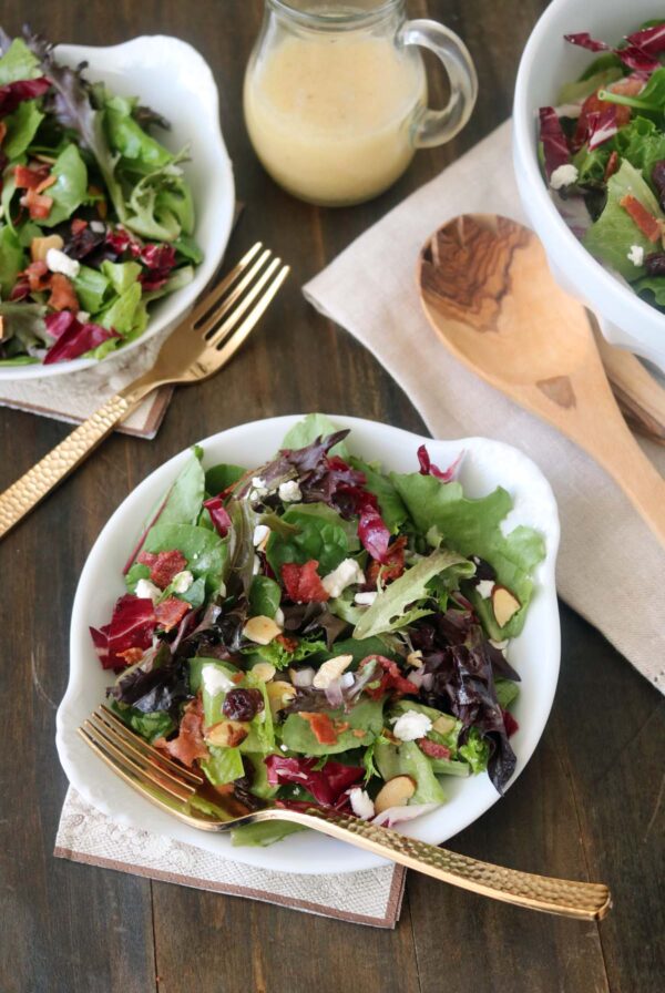 A bowl of mixed green salad with feta cheese, bacon bits, and almond slices, served with a gold fork. Two other bowls, wooden utensils, and a jar of dressing are also on a wooden table.