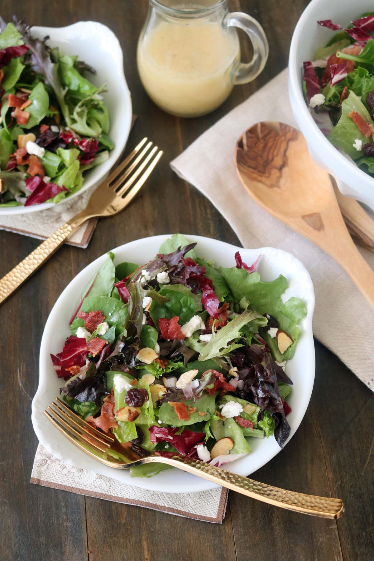A bowl of mixed green salad with feta cheese, bacon bits, and almond slices, served with a gold fork. Two other bowls, wooden utensils, and a jar of dressing are also on a wooden table.