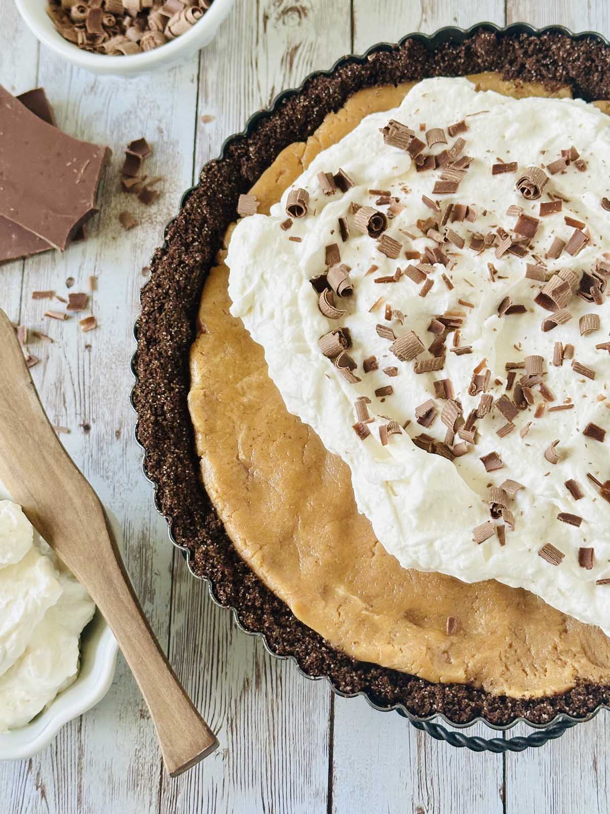 A peanut butter chocolate tart with a whipped cream topping and chocolate shavings sits in a tart pan on a wooden surface. Nearby, a bowl of whipped cream with a wooden spatula and pieces of chocolate complete the scene.