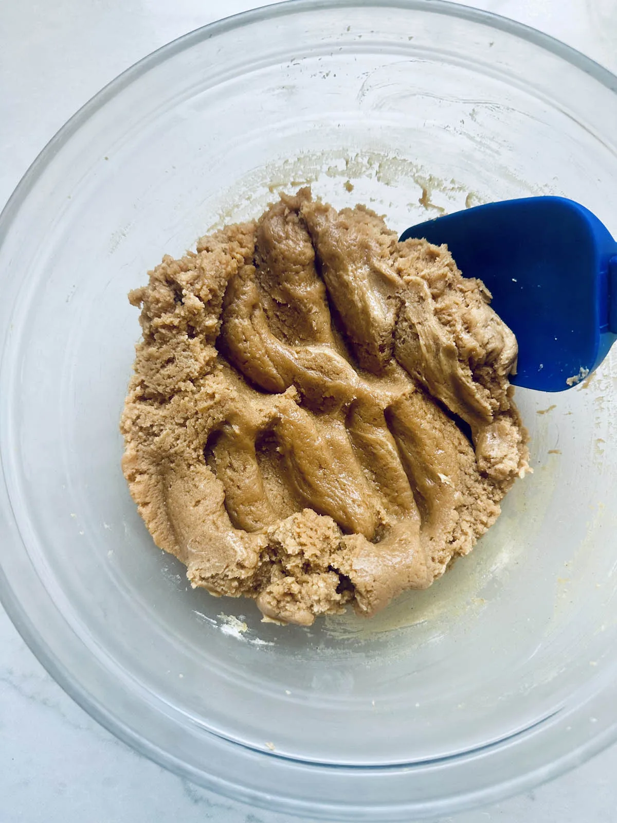 A glass bowl filled with peanut butter cookie dough is being mixed with a blue silicone spatula, the thick, creamy dough reminiscent of a base for a luscious peanut butter chocolate tart.
