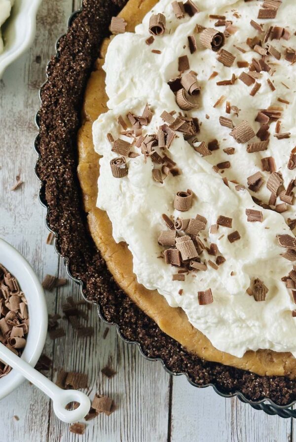 A close-up of a peanut butter chocolate tart with a chocolate crust, topped with whipped cream and chocolate shavings, sits on a rustic wooden surface. A small bowl filled with more chocolate shavings is nearby.